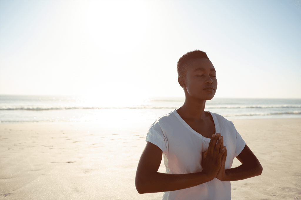 Mulher negra, cabelo curto, de camiseta branca, na praia, meditando